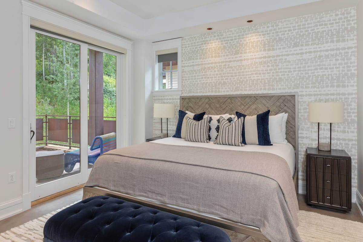 A bedroom with a large bed, a view through a sliding glass door, a headboard with a herringbone pattern, and a nightstand with a lamp.