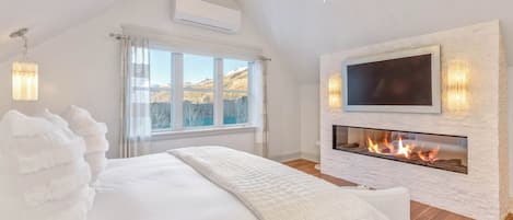 A bedroom with a large bed, a fireplace, a TV mounted on a stone wall, and a view of the outdoors.