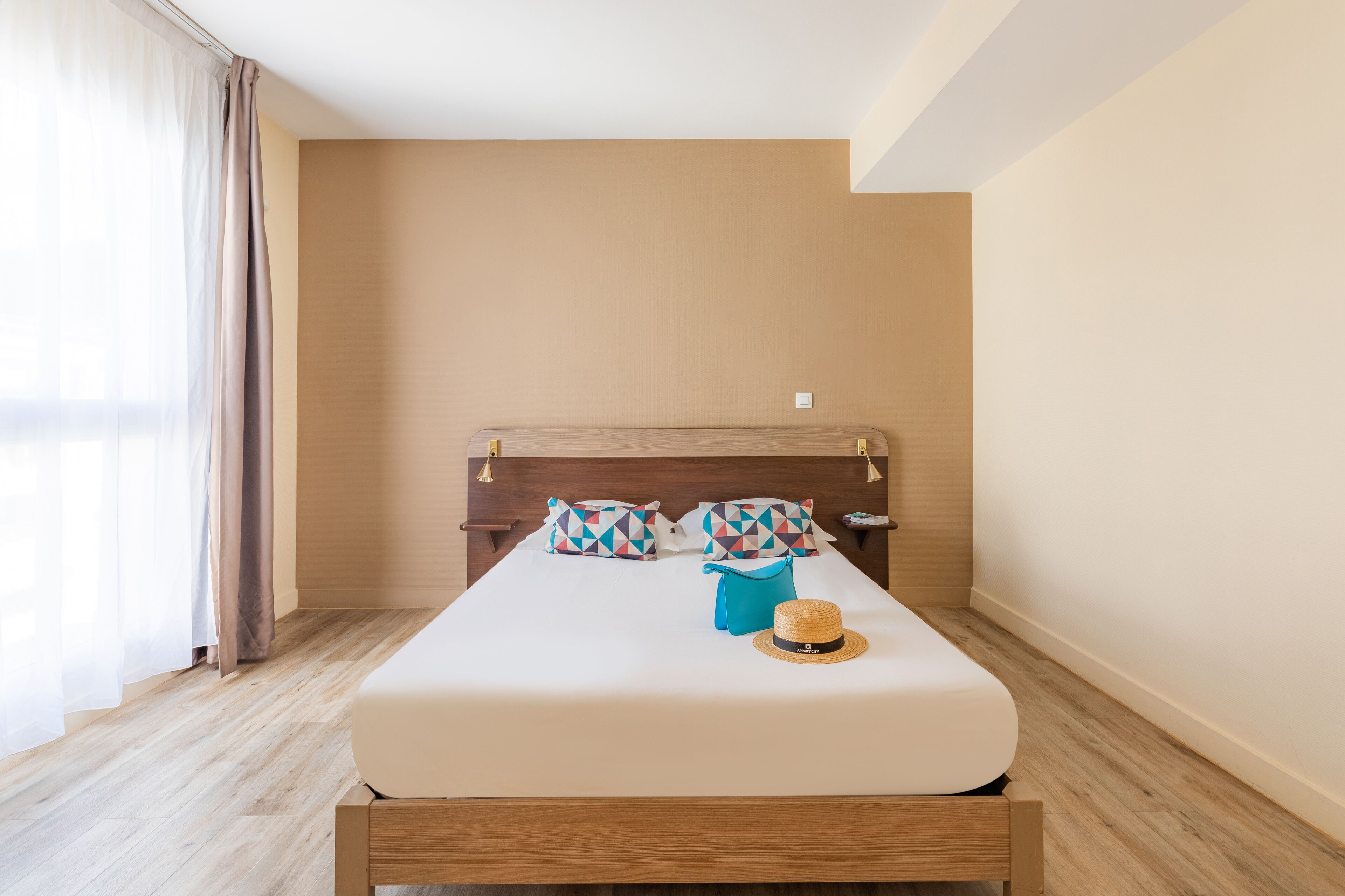 A neatly made bed with a wooden headboard, two patterned pillows, a blue bag, and a straw hat on the bed.