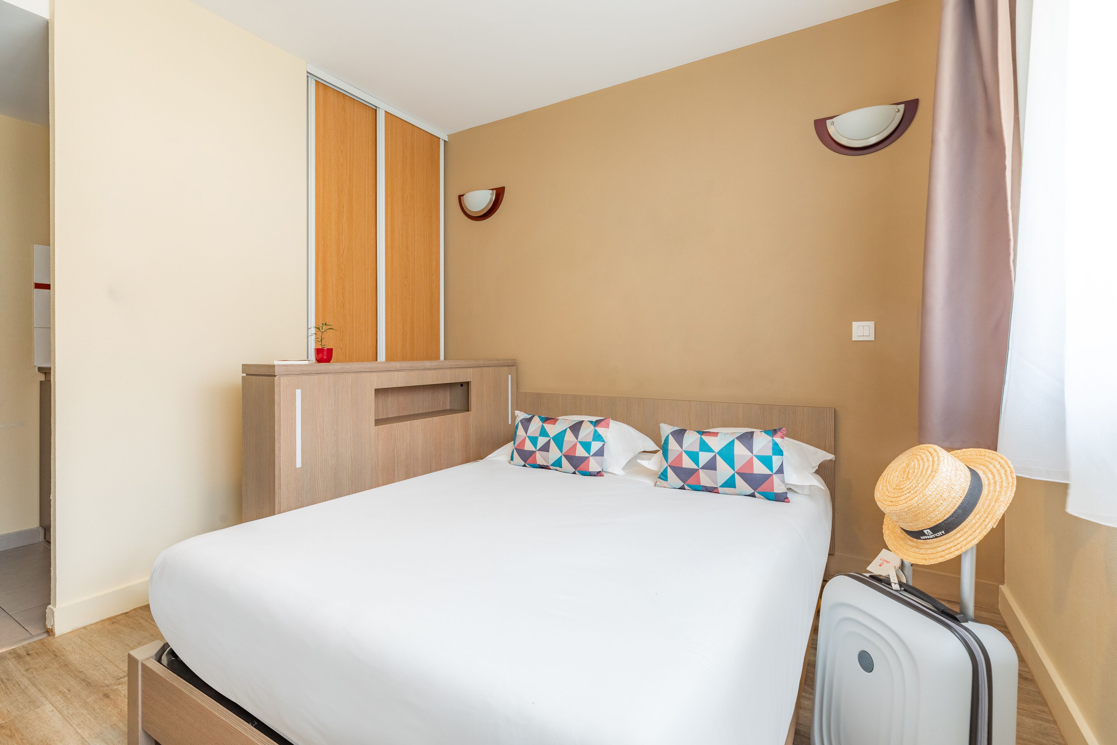 A hotel room with a bed, a wardrobe, a suitcase, and a hat on the wall.