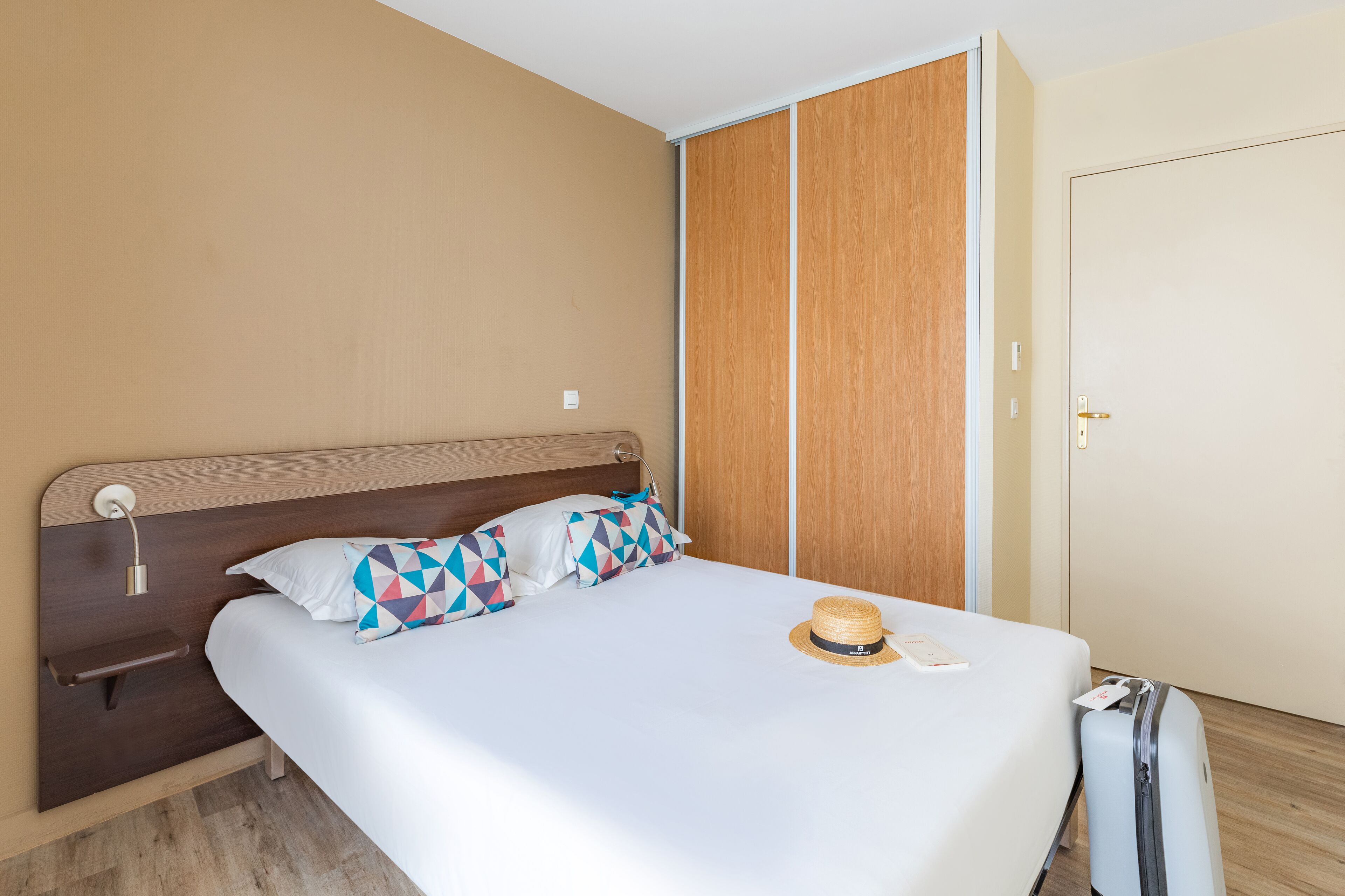 Una cama bien hecha con edredón blanco, cabecera de madera y almohadas geométricas de colores. Un sombrero de paja y una maleta están sobre la cama.