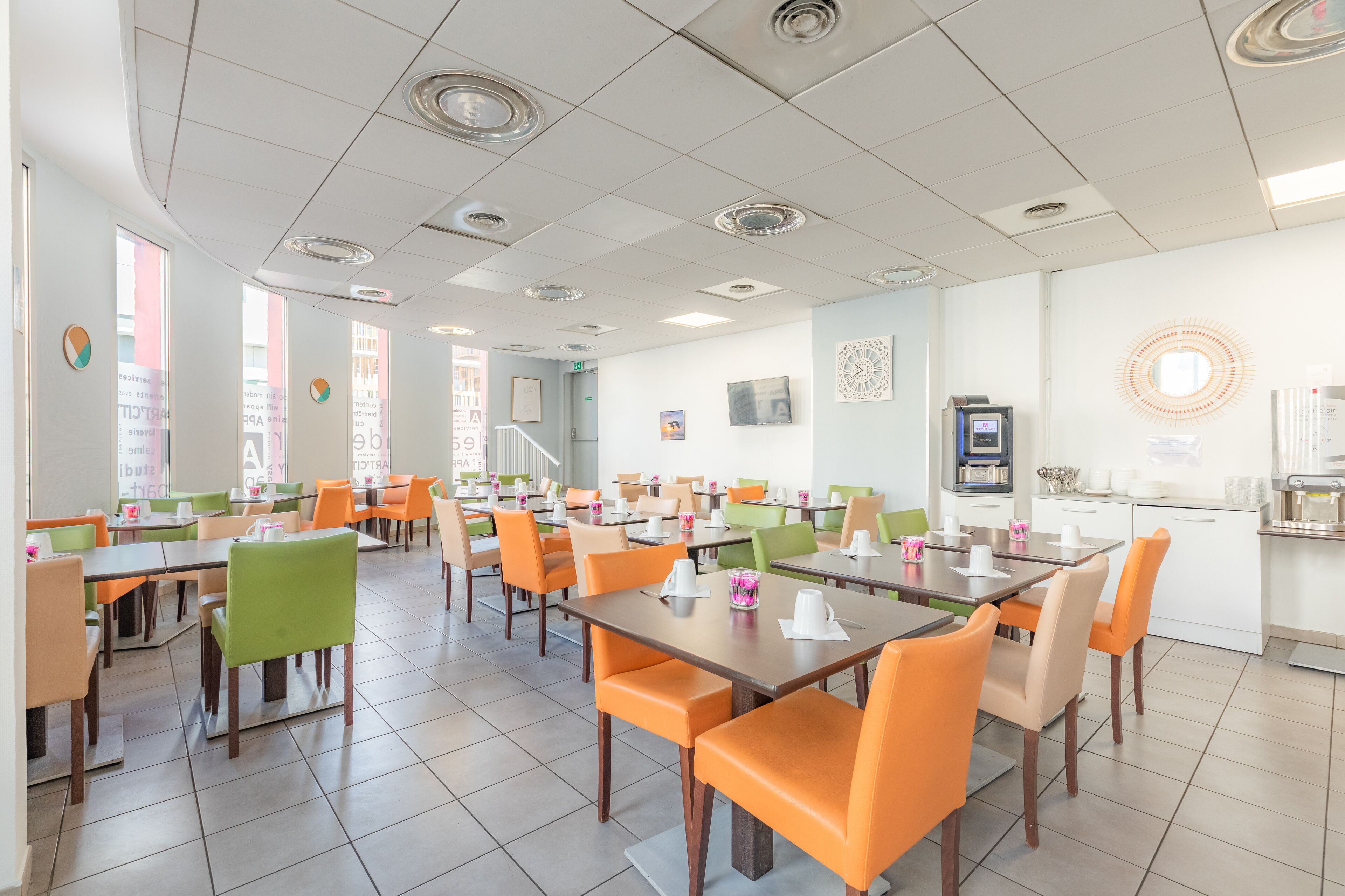 A spacious dining area with multiple tables and chairs, a kitchen area, and a staircase leading to an upper level.