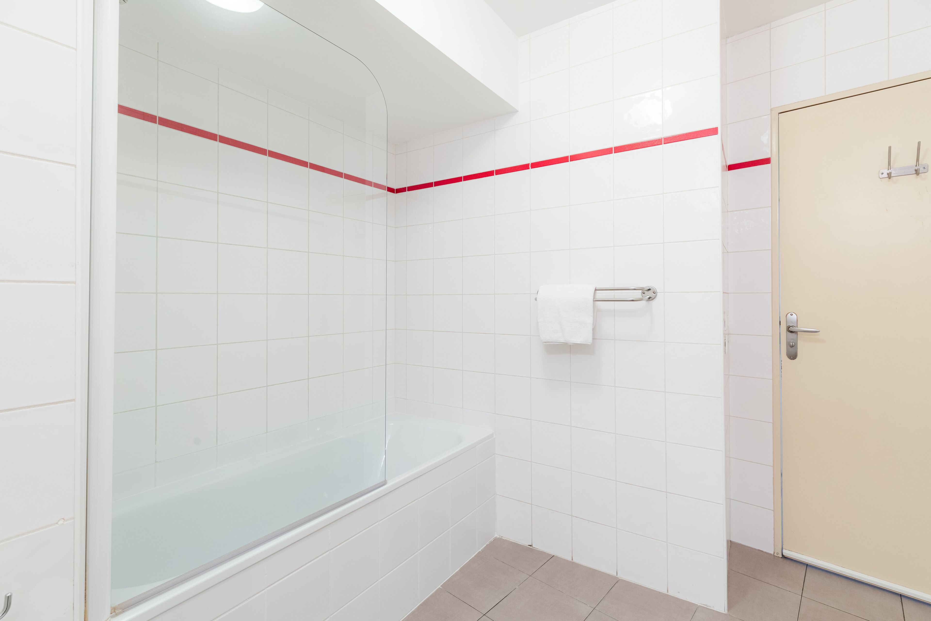 Une salle de bain avec des carreaux blancs, une douche en verre, une porte beige et une fine bande rouge décorative sur le mur.