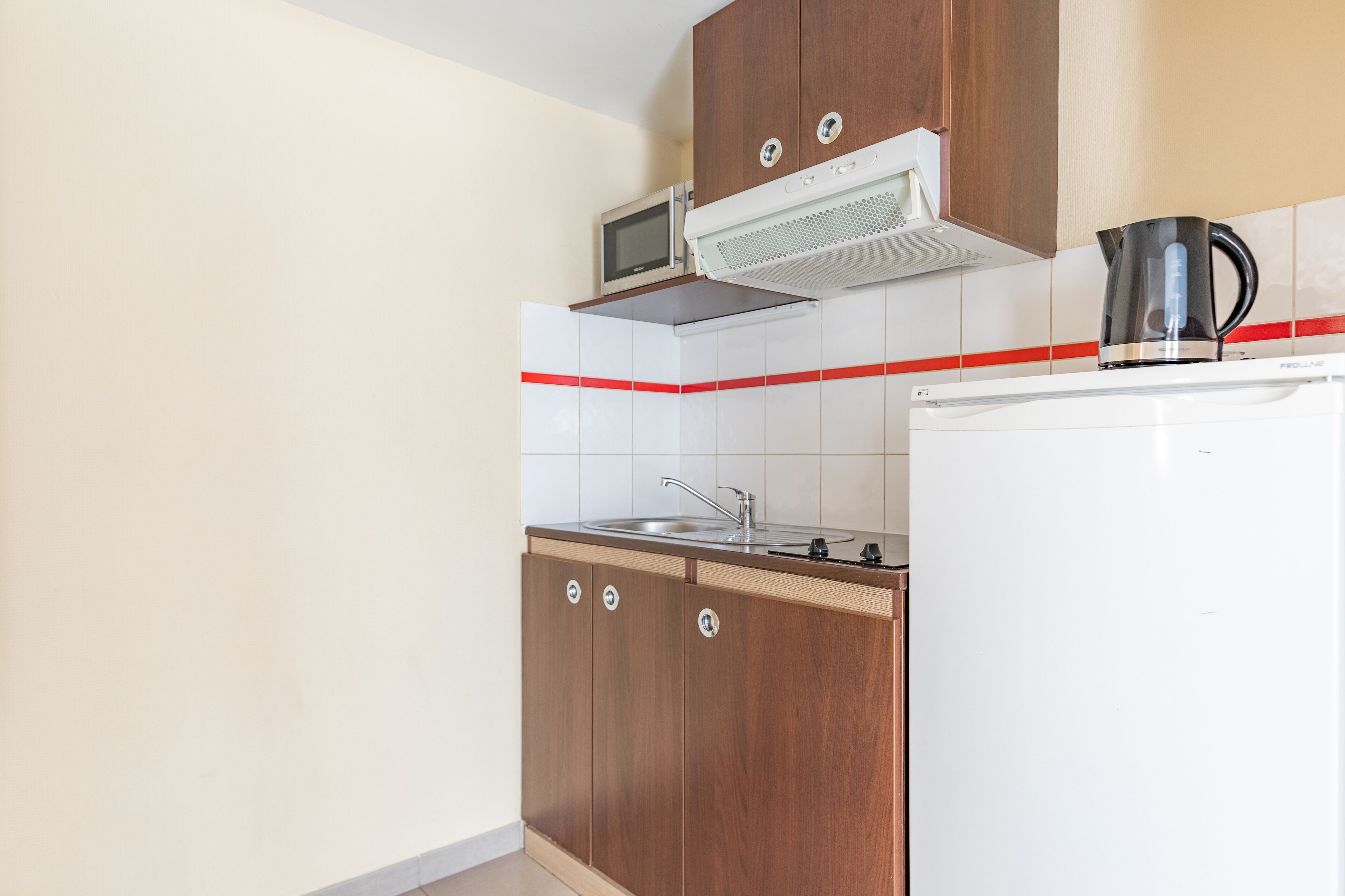 A kitchen with wooden cabinets, a microwave, a kettle, and a refrigerator.