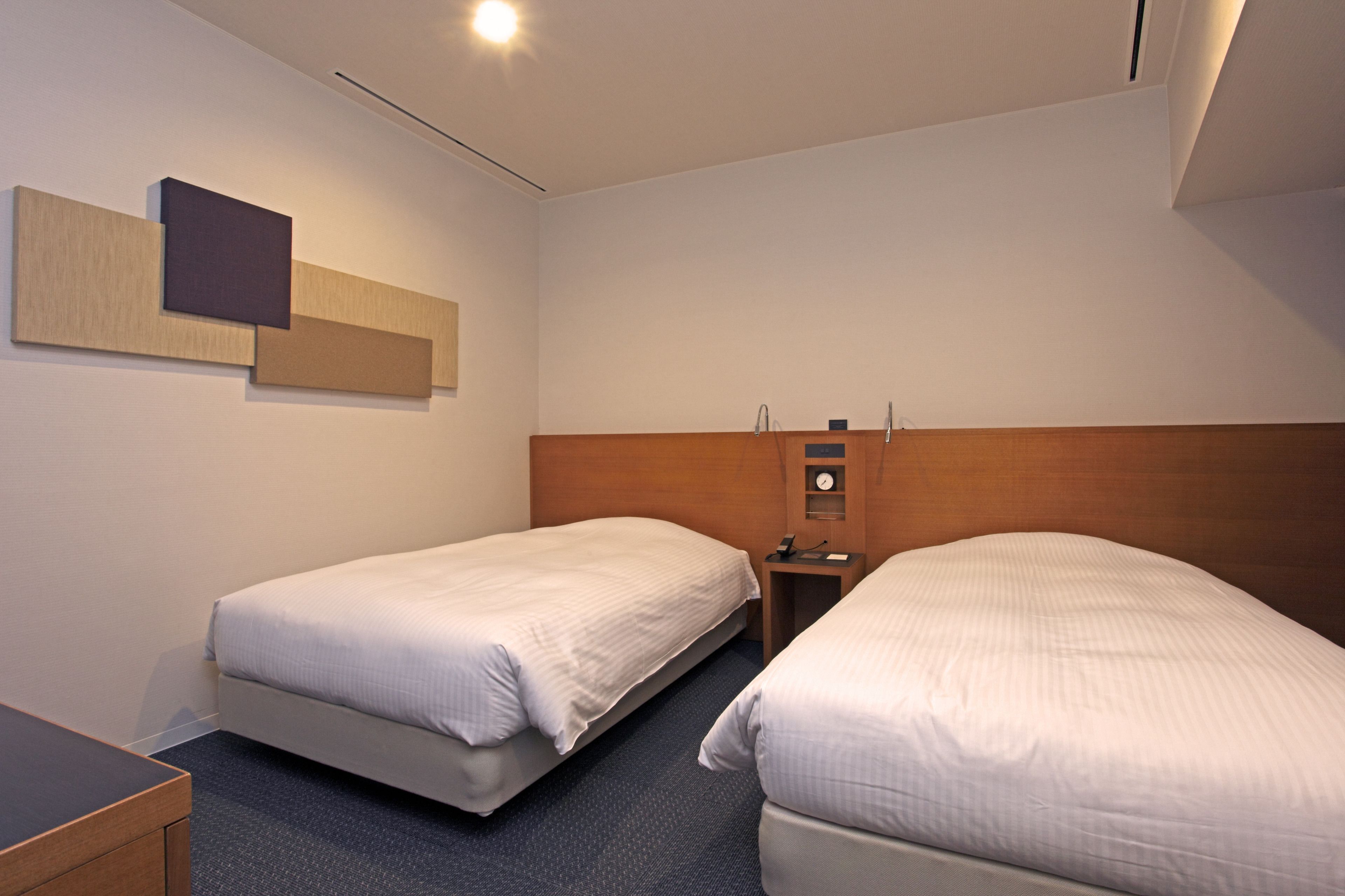 A hotel room with two single beds, a wooden headboard, a small bedside table with a lamp, and a wall-mounted clock.