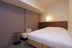 Habitación de hotel con una cama grande, una mesita redonda pequeña, una silla y una lámpara de noche.
