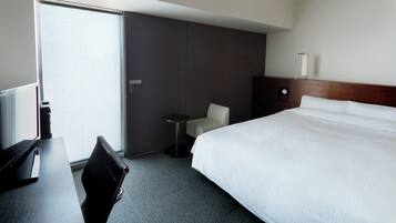 A hotel room with a large bed, a desk with a chair, a small round table, and a wall-mounted lamp.