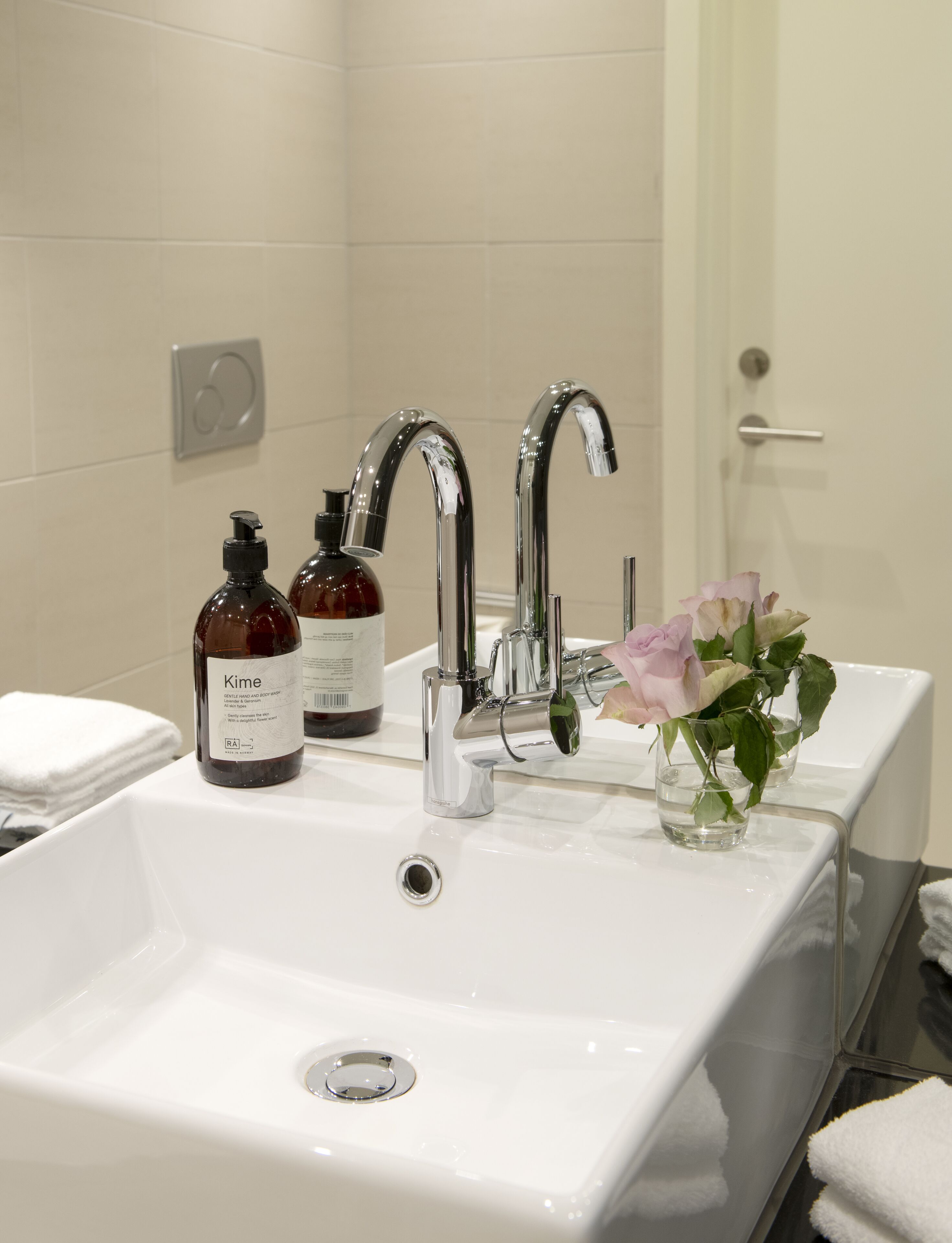 A bathroom with a white sink, two chrome faucets, and a glass vase with pink roses. There are two bottles of lotion on the sink.