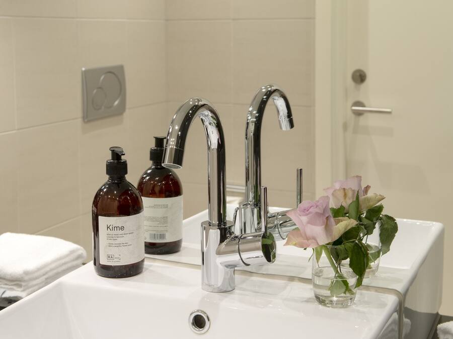 A bathroom with a white sink, two chrome faucets, and a glass vase with pink roses. There are two bottles of lotion on the sink.