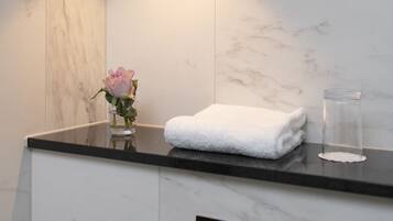 A bathroom with a marble wall, a round mirror, a white towel, a glass container, and a vase with a pink flower.