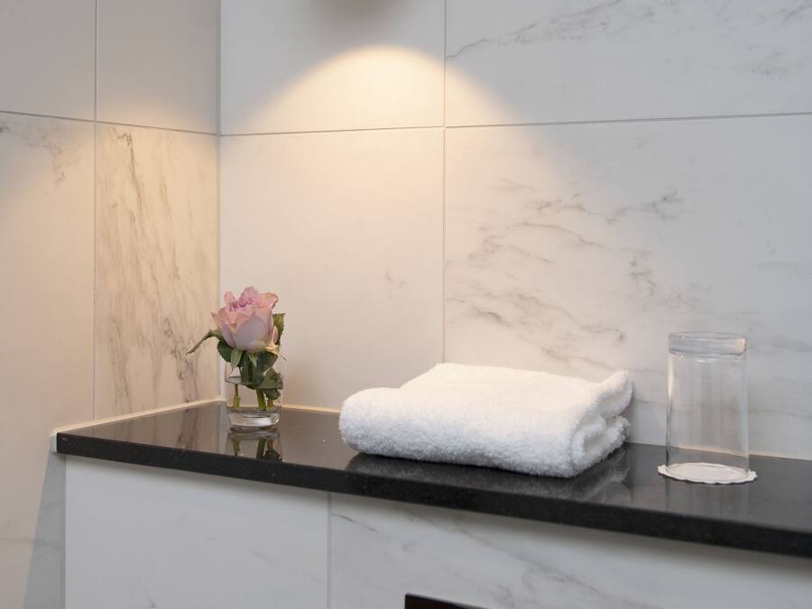 A bathroom with a marble wall, a round mirror, a white towel, a glass container, and a vase with a pink flower.