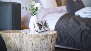 Een moderne slaapkamer met een groot bed, een nachtkastje met een vaas en een fotolijst, en een grijze gestoffeerde fauteuil.