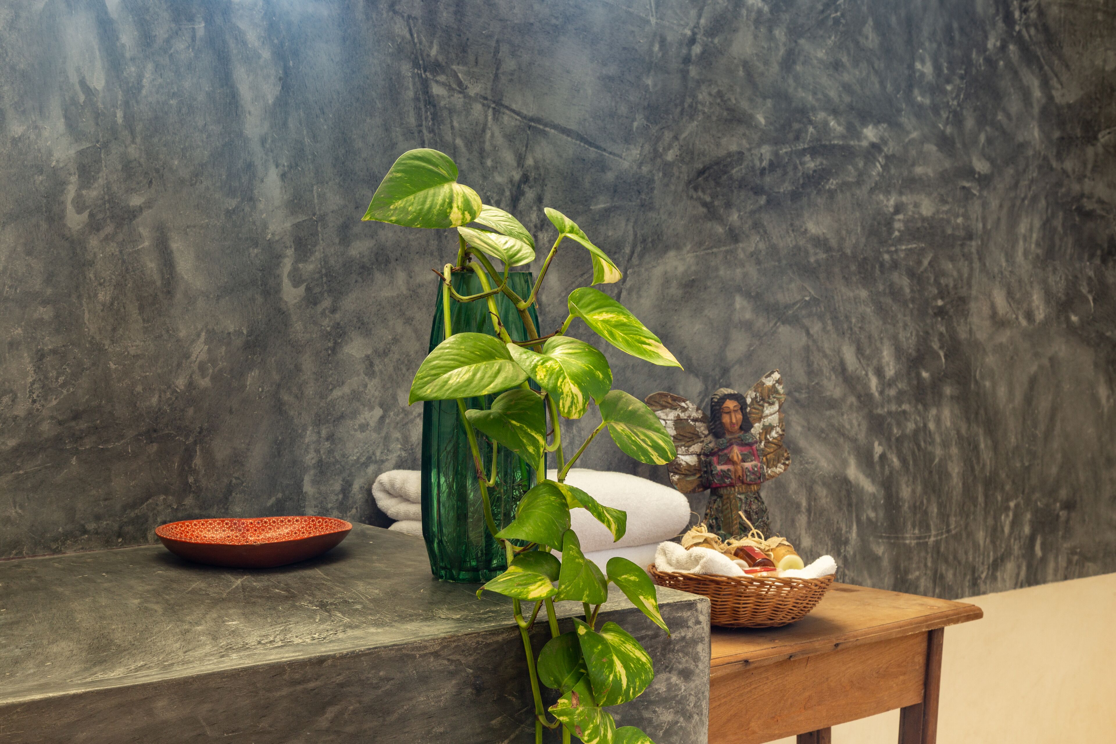 A bathroom with a marble countertop, a green plant, a red bowl, a green vase, a wicker basket with towels, and a statue.