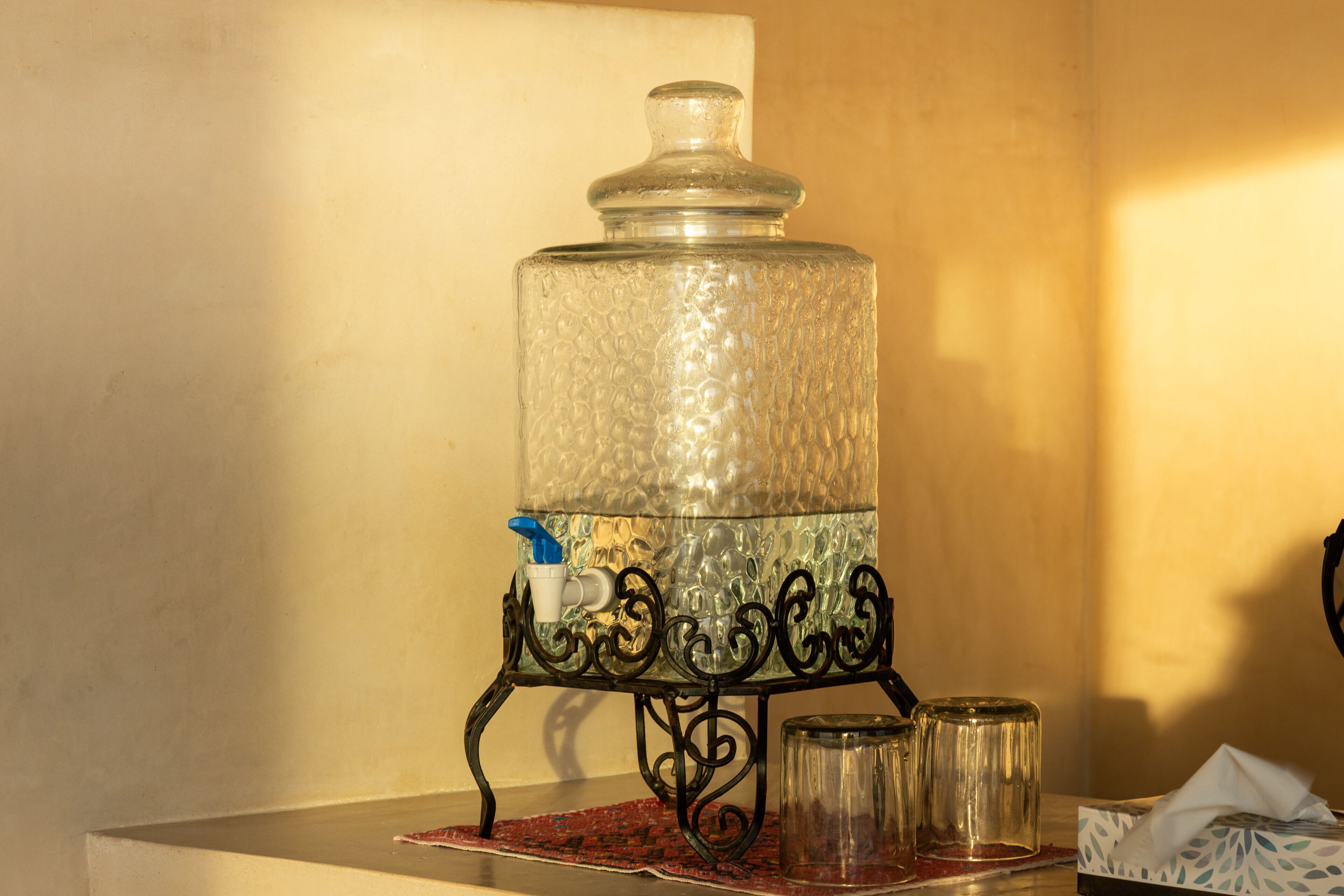 A water dispenser on a metal stand with a glass container, a bottle, and a tissue box.