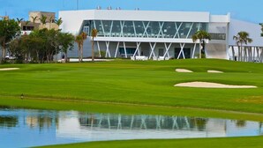 Um campo de golfe com um edifício moderno ao fundo, um lago e palmeiras.