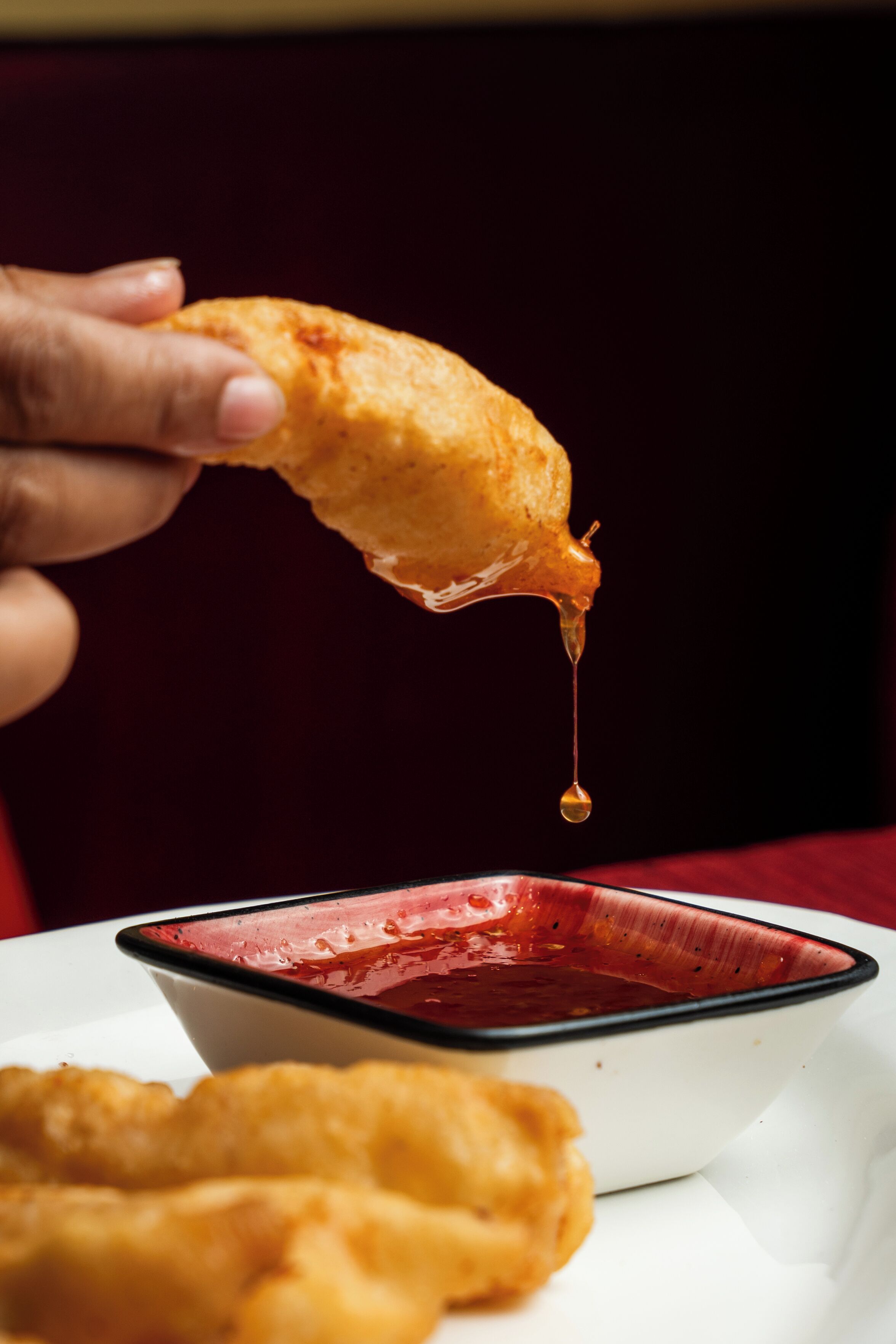 A hand holding a fried item with sauce dripping, next to a bowl of sauce.