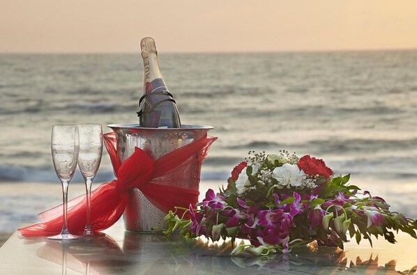 A bottle of champagne in a metal bucket with a red ribbon, two empty glasses, and a bouquet of flowers on a table by the ocean.