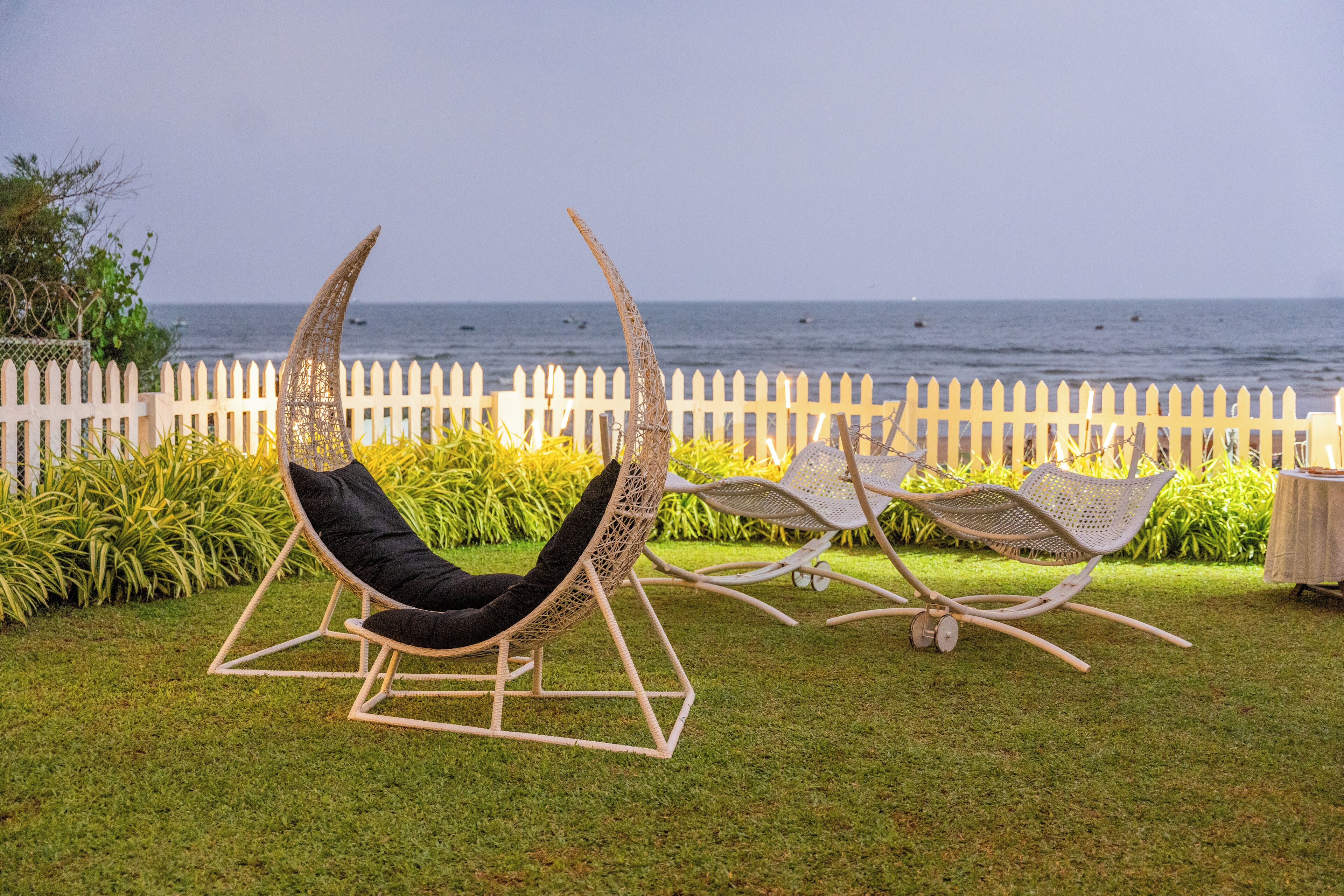 Two wicker lounge chairs with black cushions in a grassy area with a white picket fence and a view of the sea.