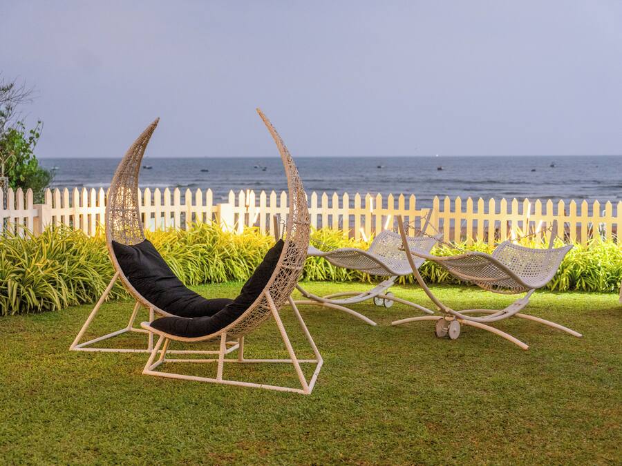 Dos sillones de mimbre con cojines negros en un área con césped, una valla de piquete blanca y vista al mar.
