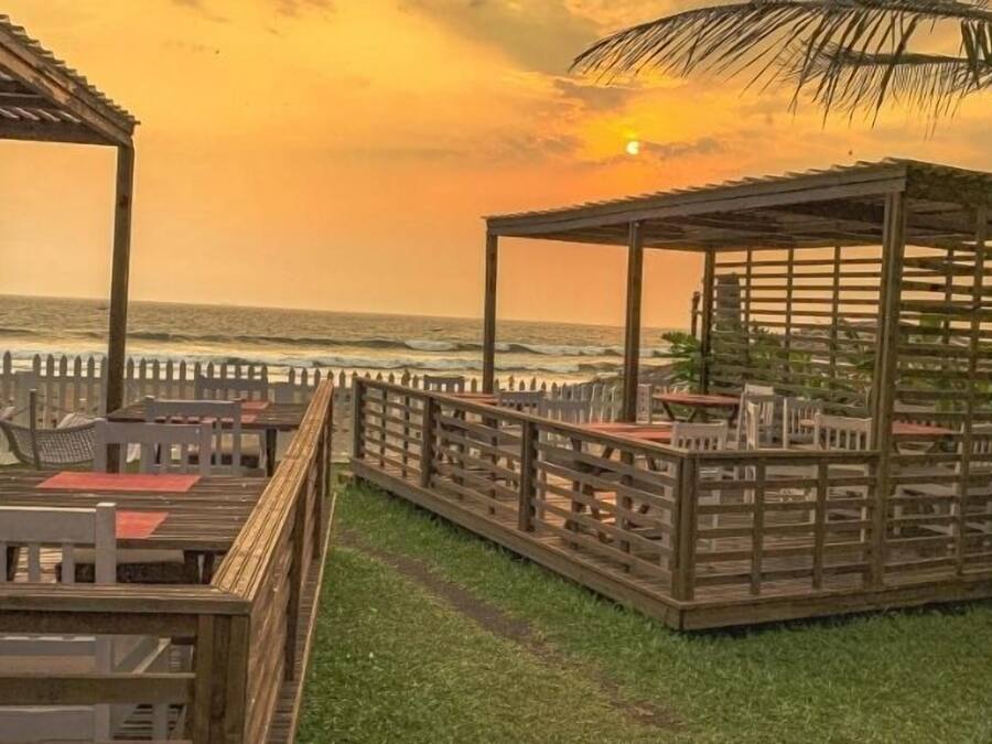 Um deck de madeira com mesas e cadeiras, com vista para o oceano ao pôr do sol.