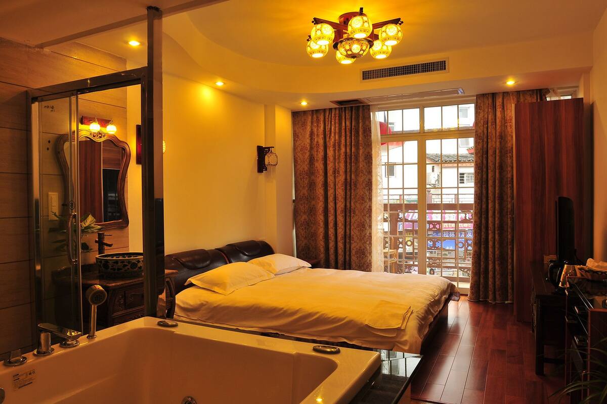 A hotel room with a large bed, a bathtub, a mirror, and a view of a traditional building through the window.