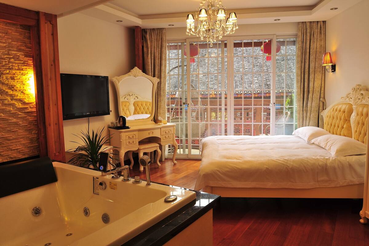 A bedroom with a large bed, a vanity with a mirror, a bathtub, and a television on the wall.