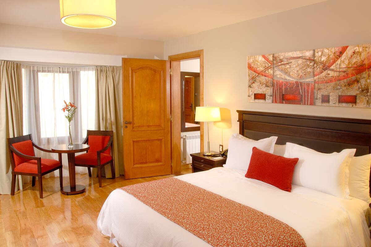 Een hotelkamer met een groot bed, een ronde tafel met stoelen, een lamp en een schilderij aan de muur.