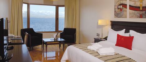 A hotel room with a large bed, a desk, two chairs, a view of the sea, and artwork on the wall.
