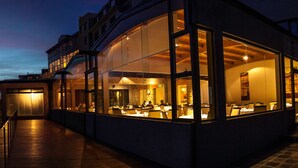 Un bâtiment moderne doté de grandes fenêtres en verre, d’une terrasse en bois et d’un éclairage intérieur.