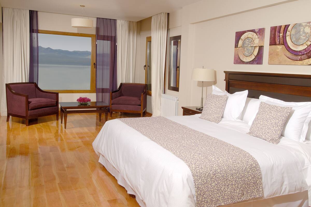 Hotelkamer met een groot bed, twee fauteuils, een tafeltje met een bloem, uitzicht op bergen door een raam, en twee abstracte schilderijen aan de muur.
