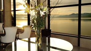 Une table ronde en verre, ornée d’un vase de fleurs, deux chaises blanches et une vue sur un lac paisible et des montagnes, le tout visible à travers de grandes fenêtres.