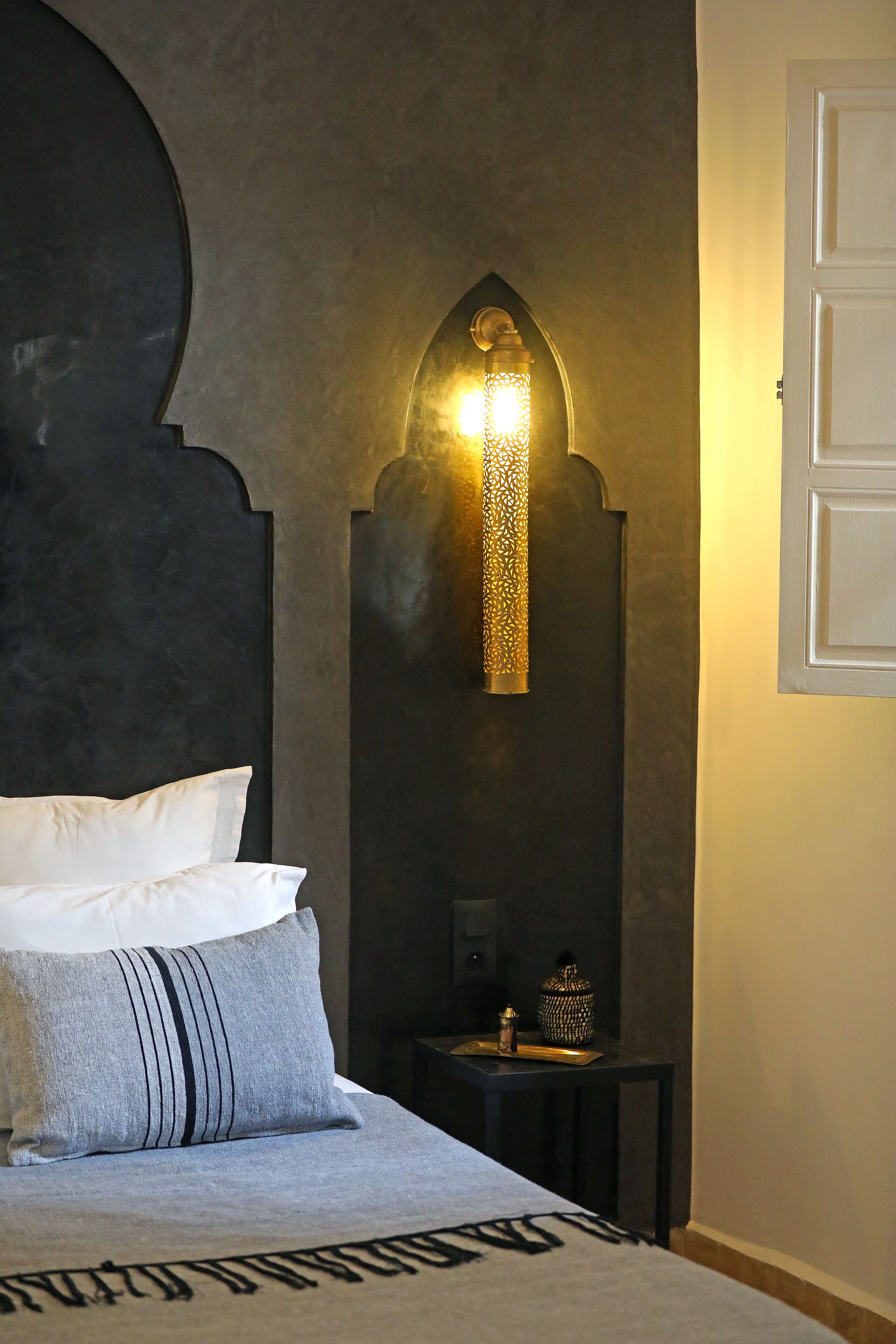 A bedroom with a dark headboard, a bed with white and striped pillows, a small bedside table with a decorative item, and a wall-mounted light fixture.