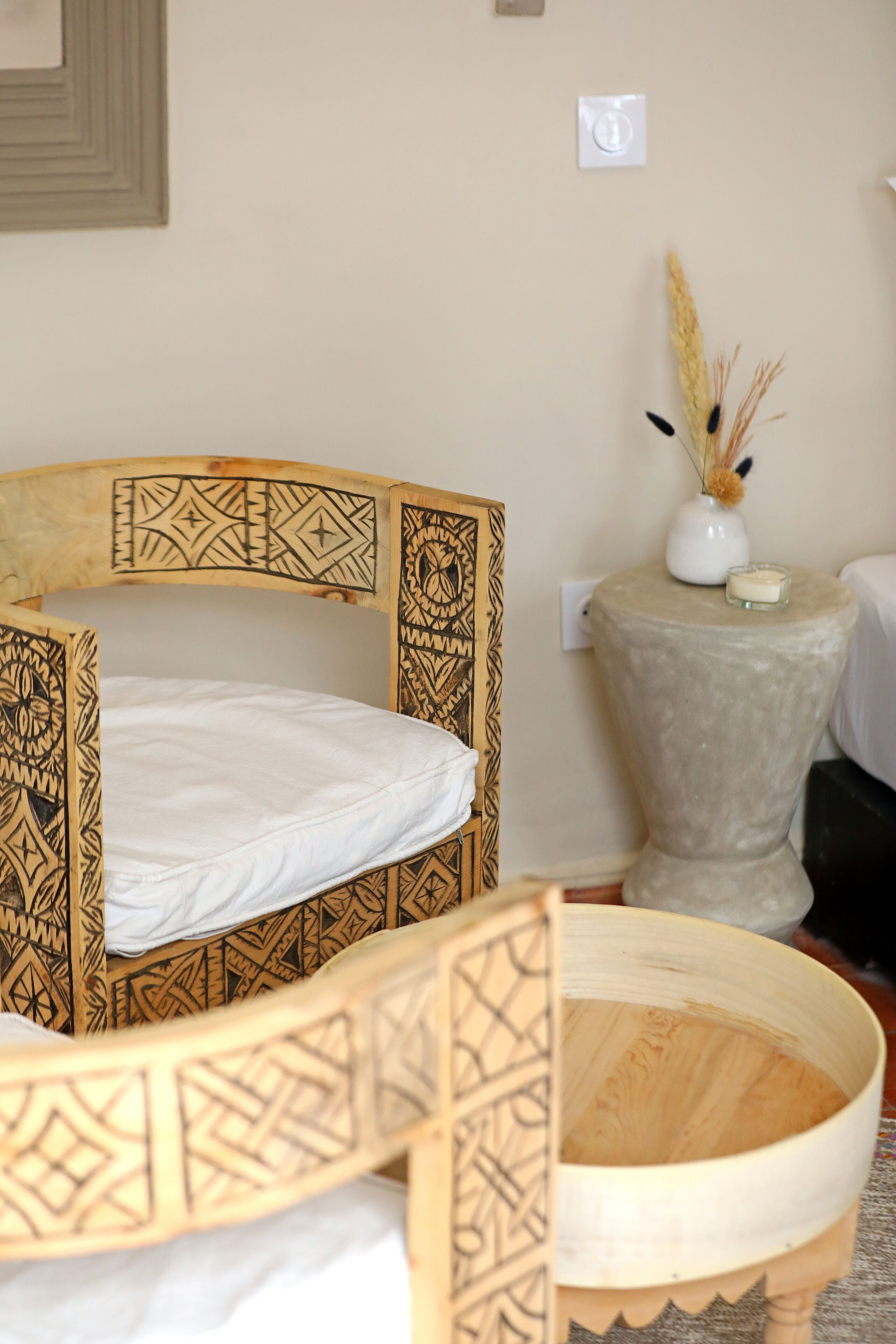 A bed with intricate wooden carvings, a bedside table with a candle and decorative items, and a wall-mounted light switch.