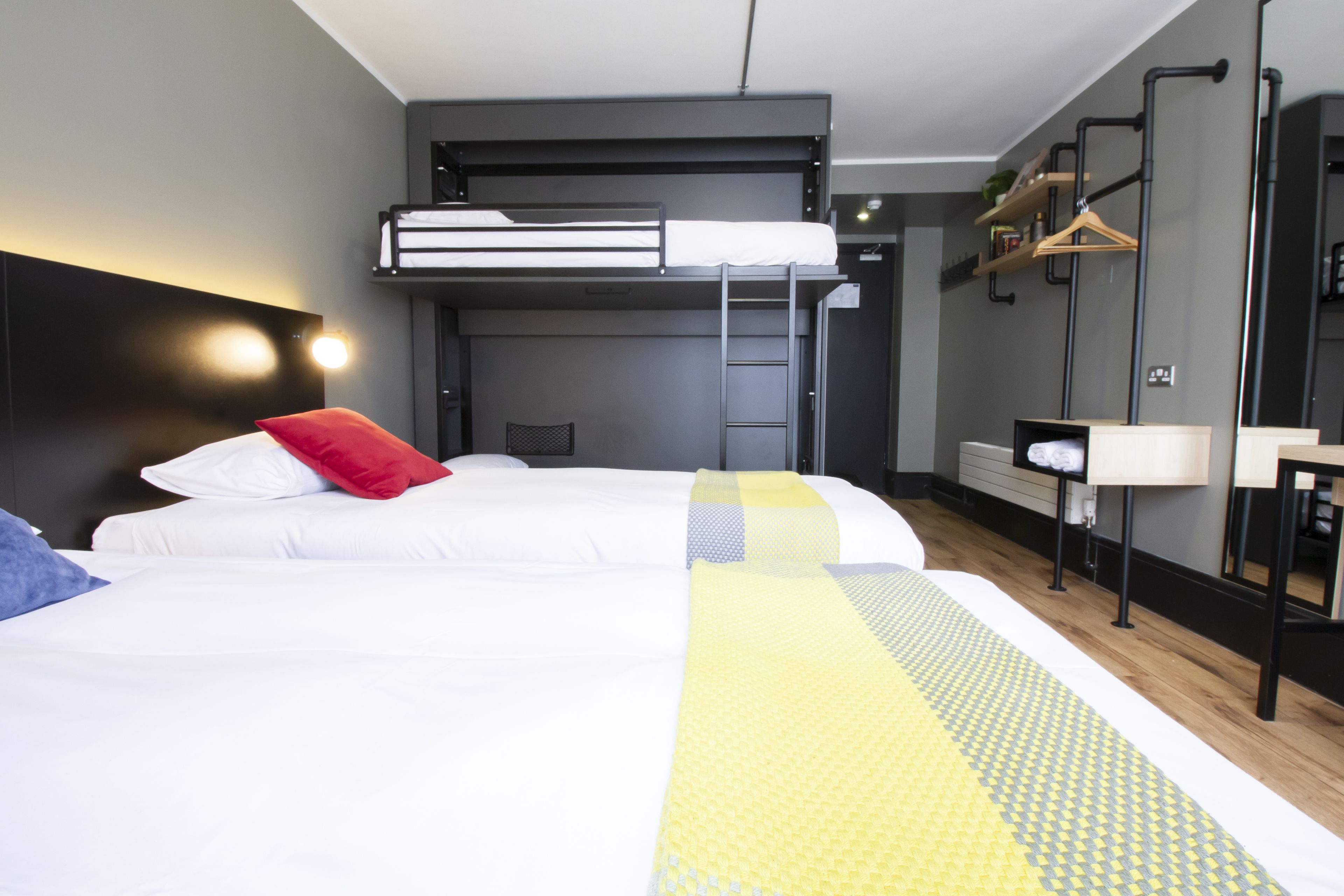 Hotellihuone, jossa on kerrossänky, työpöytä tuolilla, peili ja hylly, jossa on tavaroita.