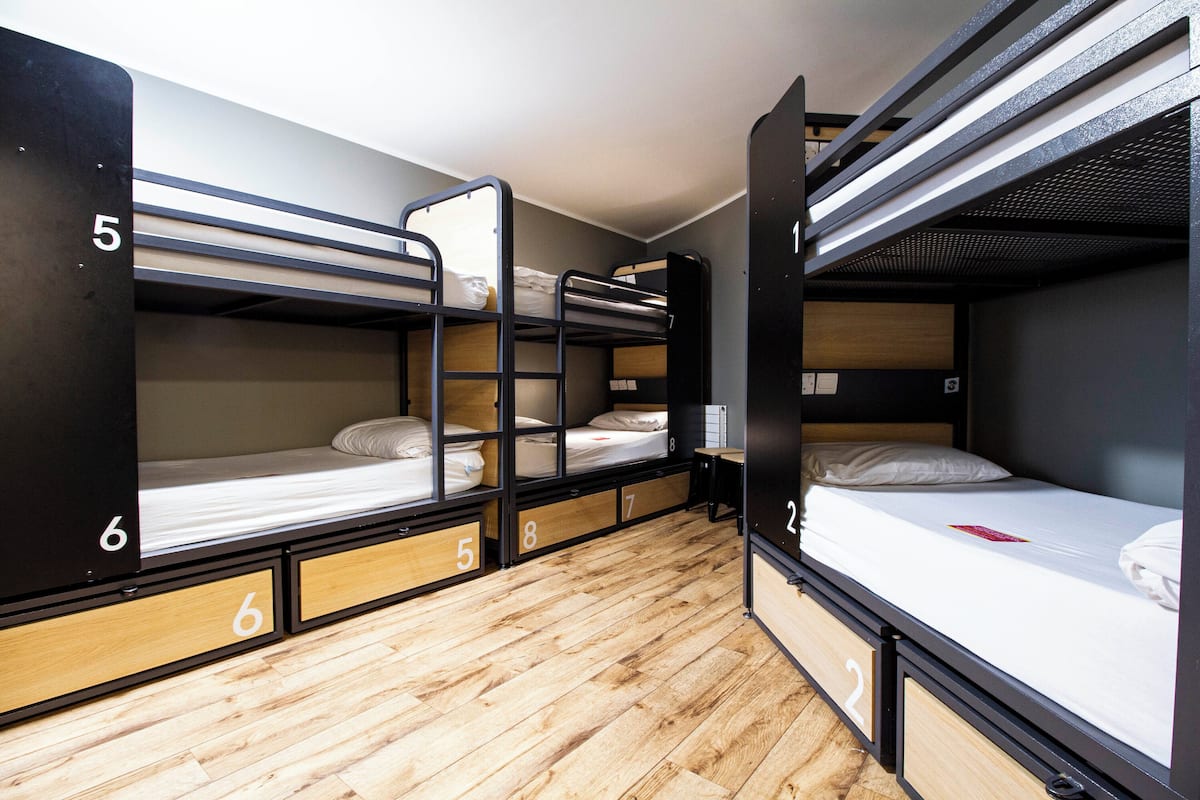 A dormitory-style room with bunk beds numbered 2 to 8, wooden flooring, and built-in storage drawers.