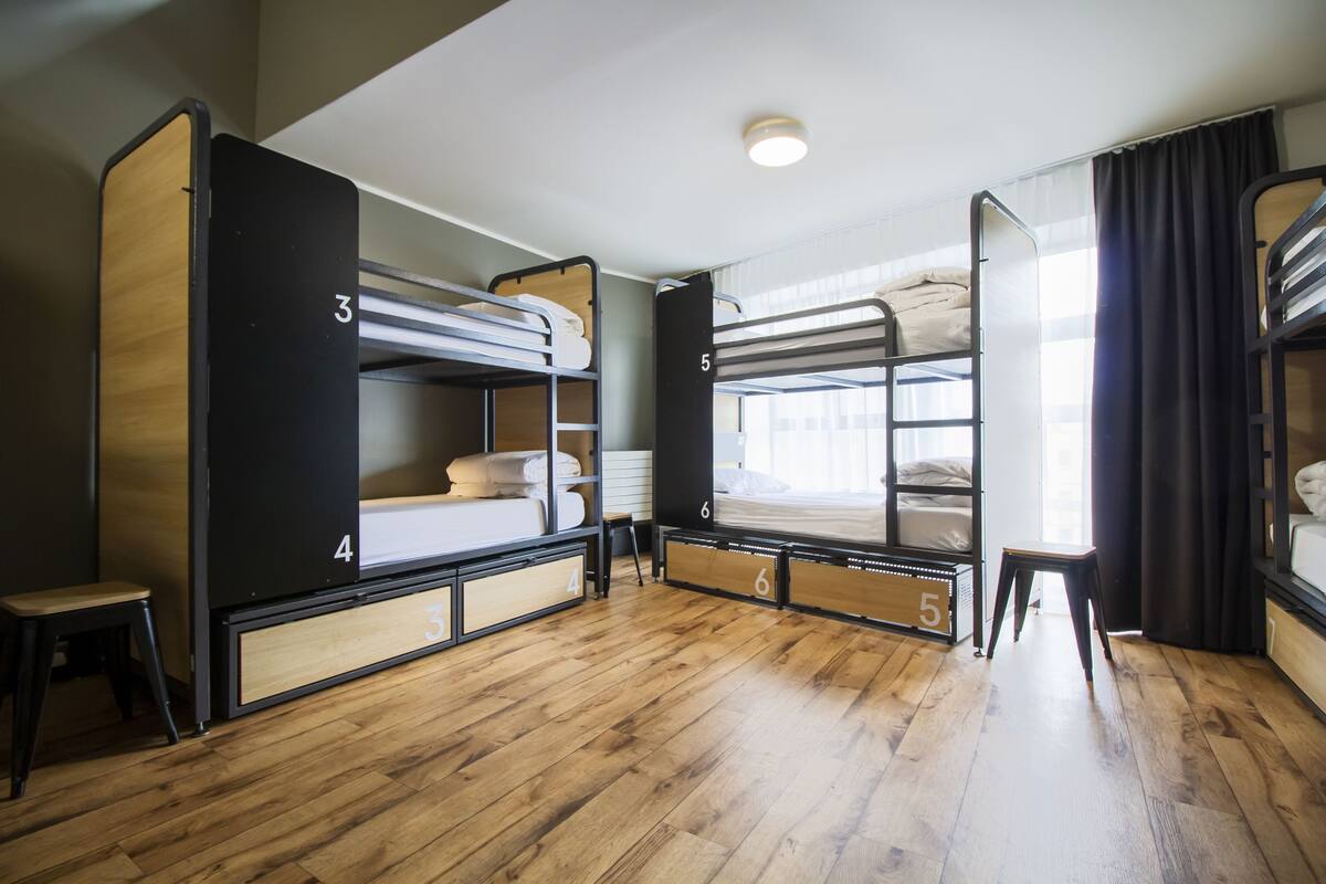 A dormitory room with bunk beds, wooden flooring, and a small stool.
