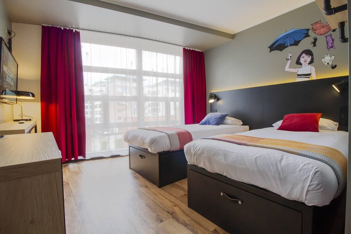 A hotel room with two beds, a desk, a chair, and a large window with red curtains.