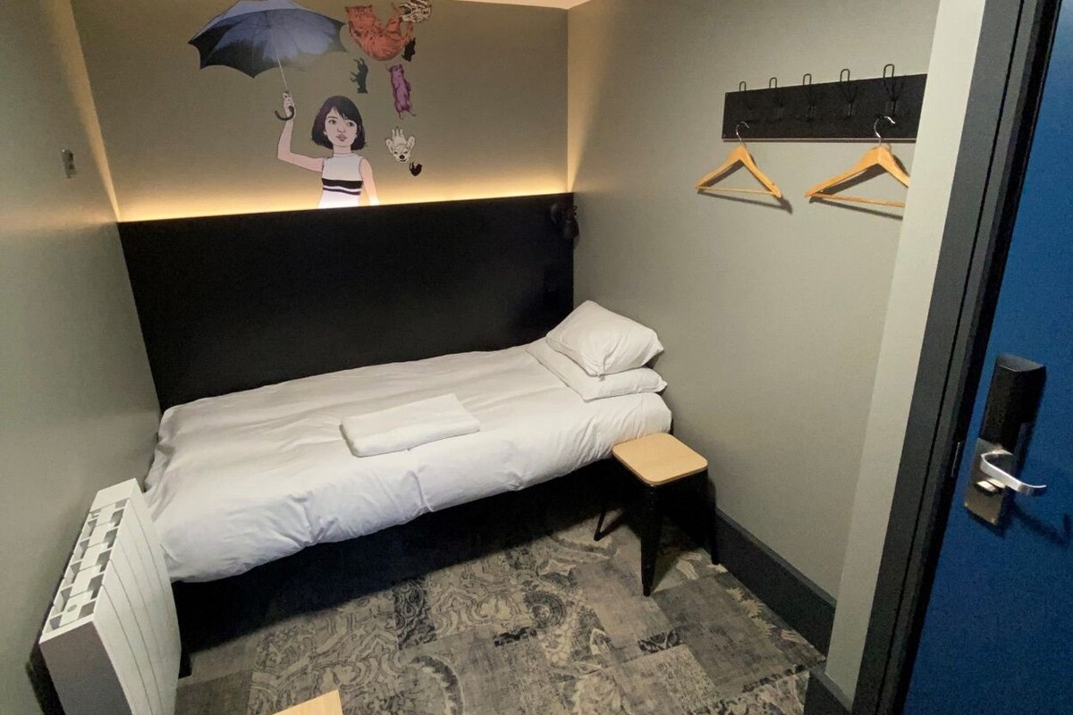 Una cama individual con cabecera, una mesita de noche pequeña, un perchero en la pared y una alfombra estampada en el suelo.
