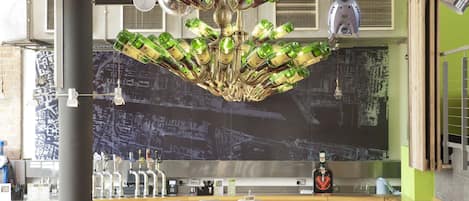 A modern restaurant interior with high ceilings, exposed ductwork, and a unique chandelier made of green bottles. Tables and chairs are arranged for dining.