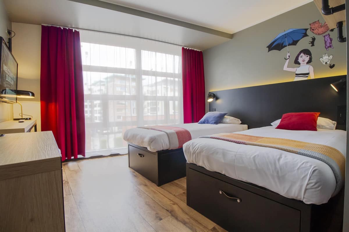A hotel room with two beds, a desk, a chair, a lamp, and a large window with red curtains.