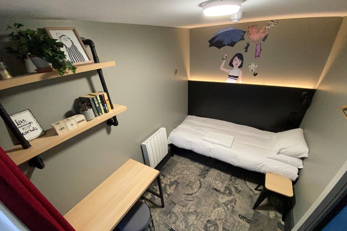 A small, neatly arranged bedroom with a single bed, a desk, and shelves with books and decorative items.