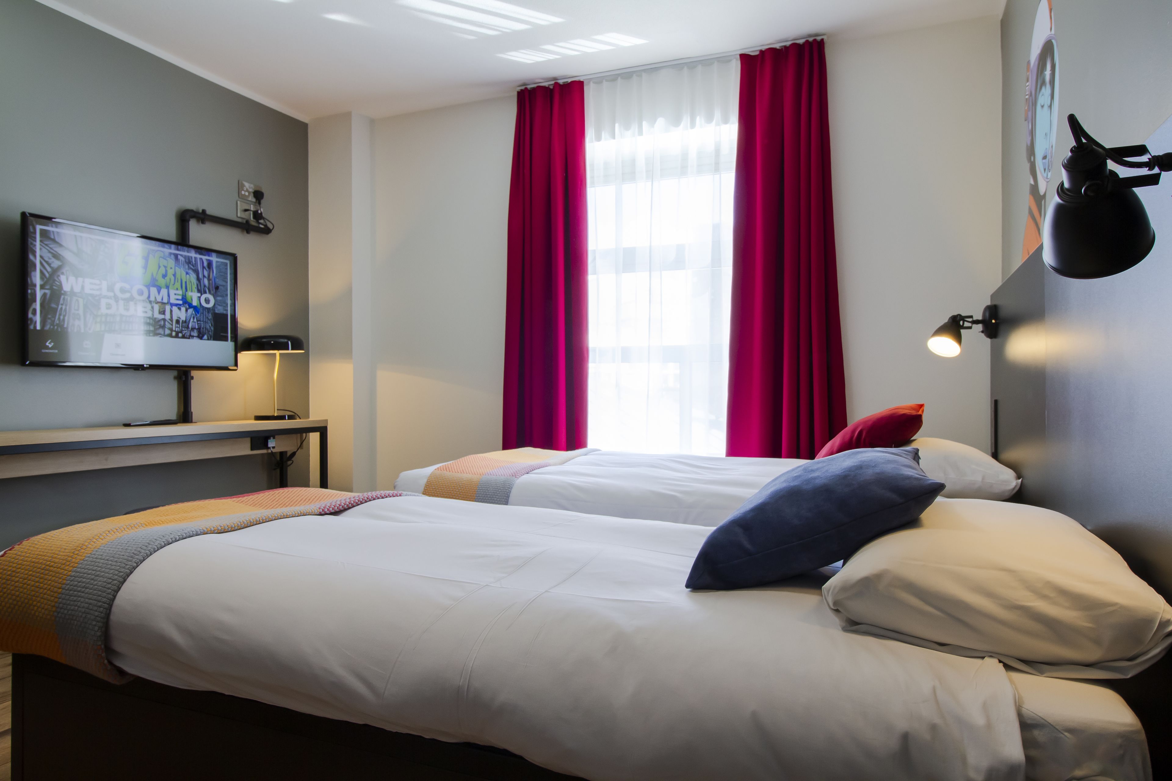 A hotel room with two beds, a flat-screen TV, a desk with a lamp, and a window with red curtains.