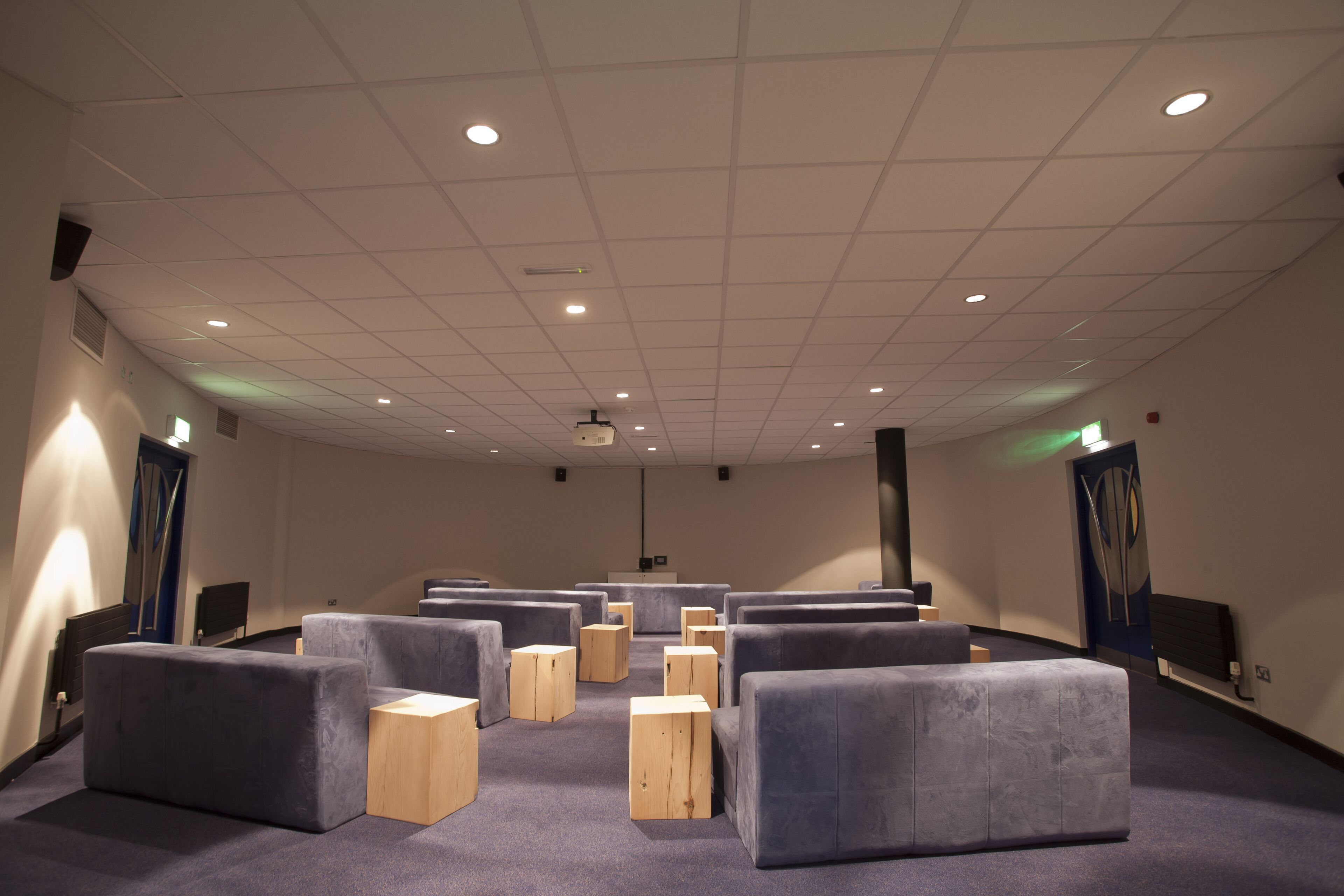 A conference room with rows of grey upholstered chairs and wooden stools, a projector screen, and a white ceiling with recessed lighting.