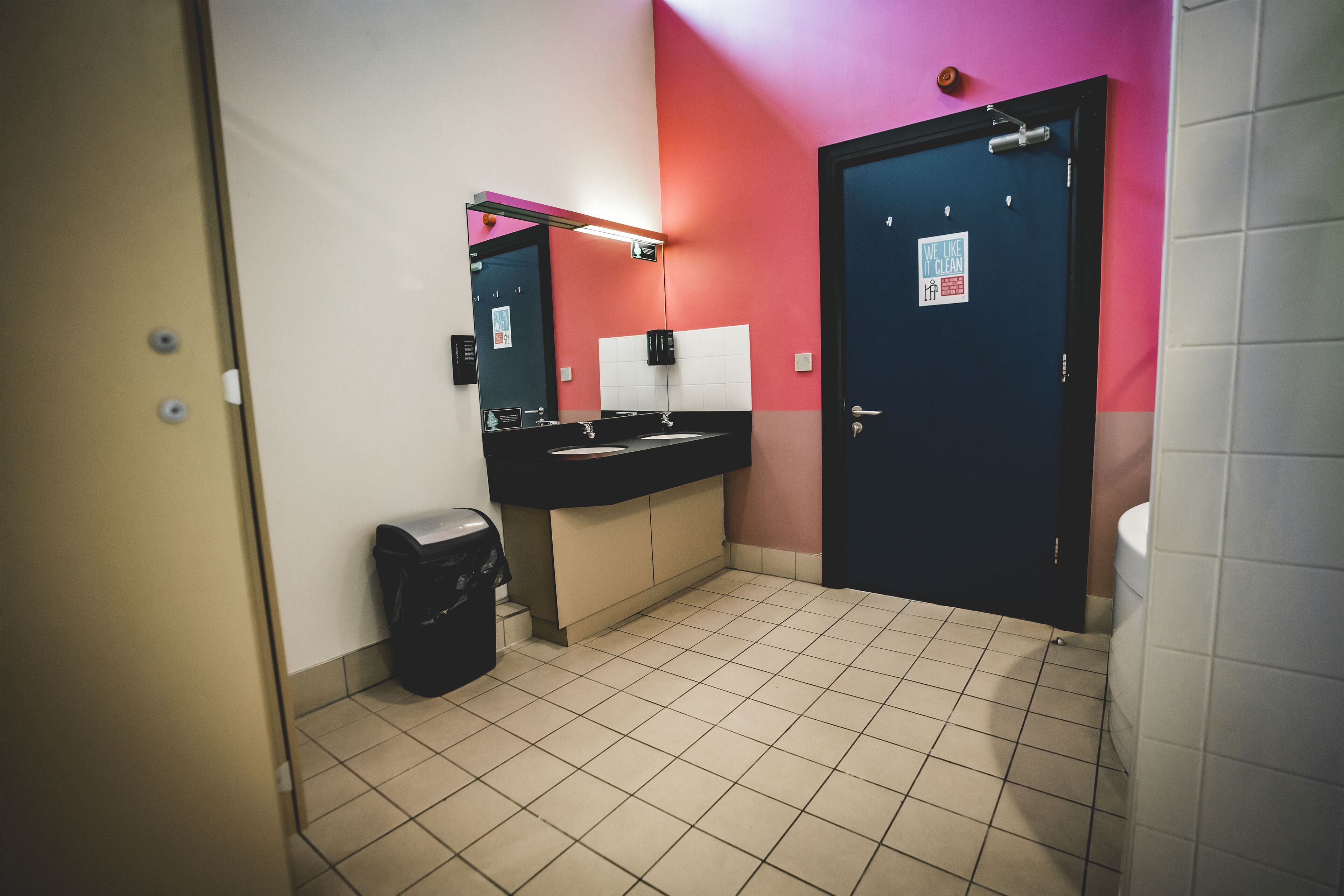 Un baño con un lavamanos negro, un cubo de basura, un espejo y una puerta con un letrero.
