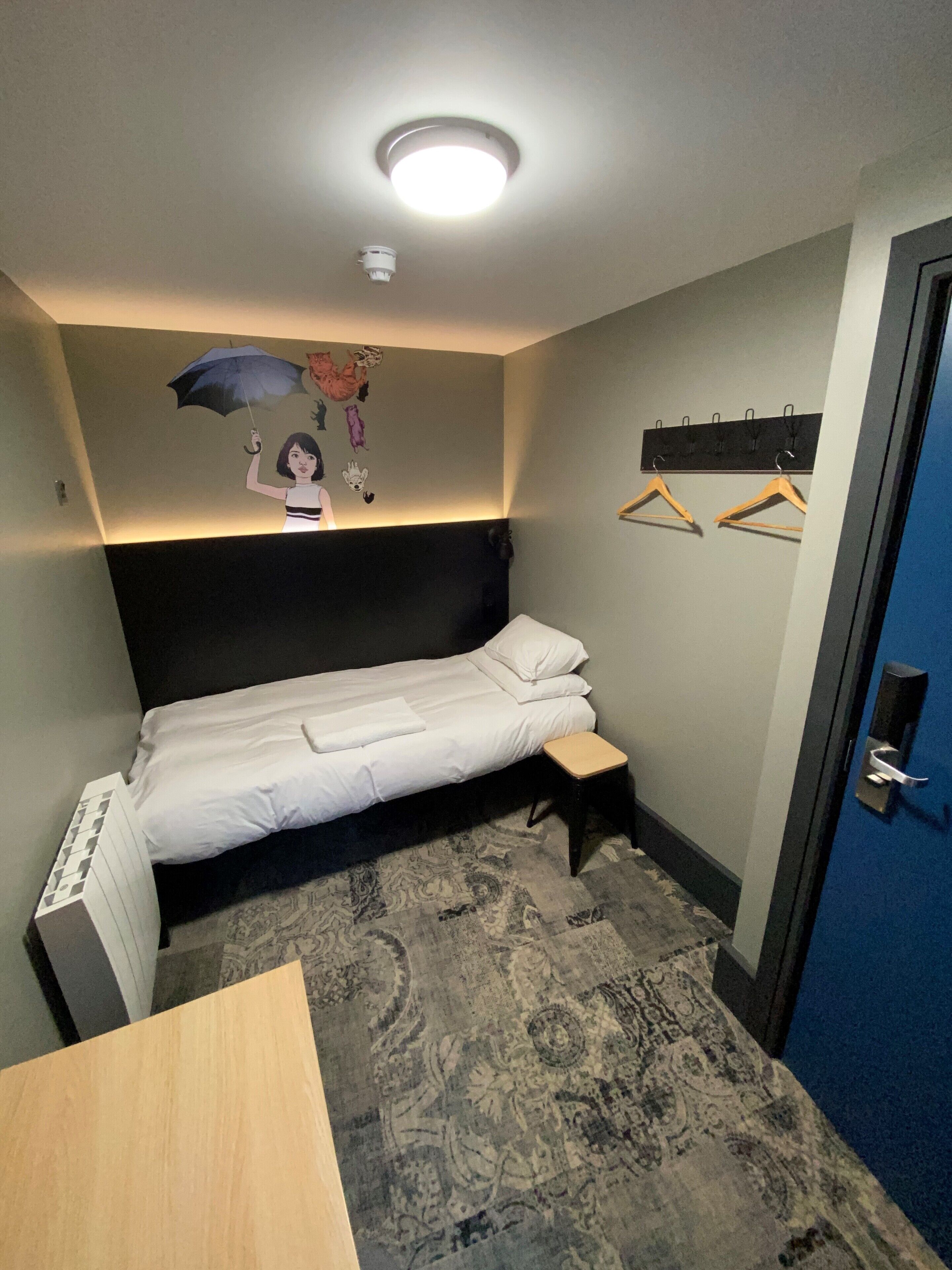 A small, neatly arranged room with a single bed, a bedside table, a wall-mounted coat rack, and a door.
