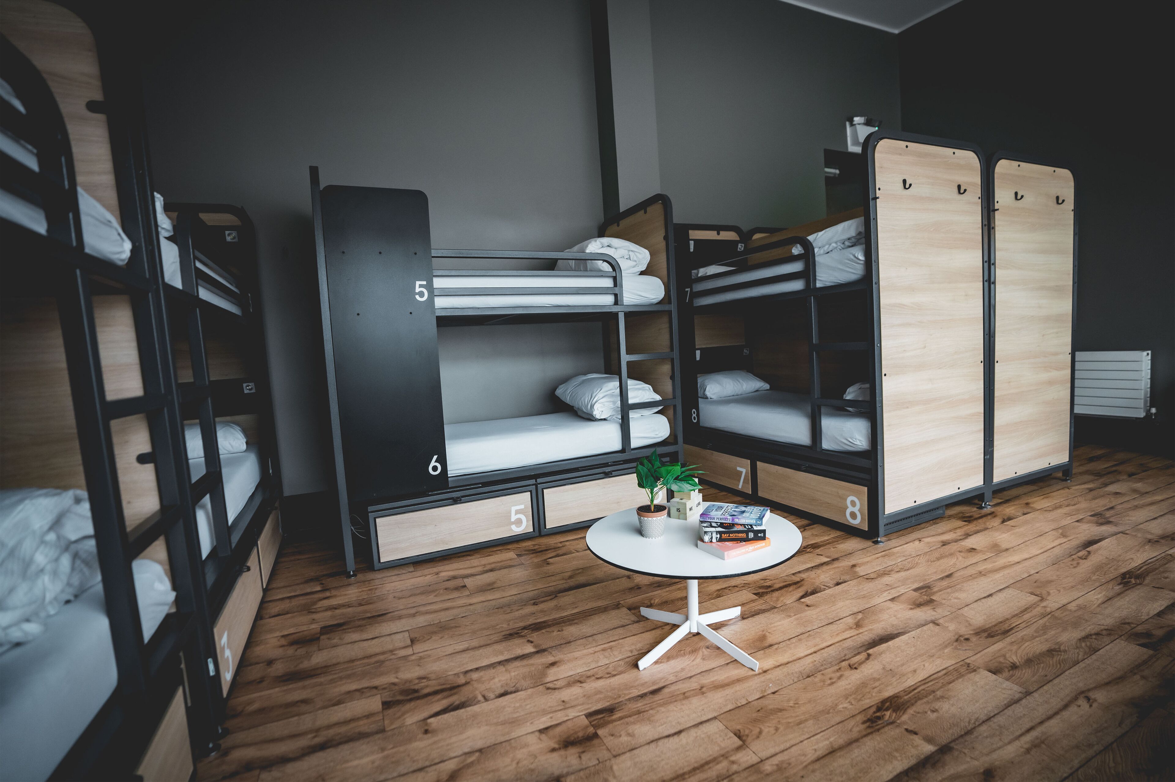 A dormitory room with bunk beds numbered 3 to 8, a small round table with a plant, and a radiator.