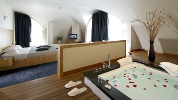 Habitación de hotel con cama, escritorio, televisor, bañera con pétalos de rosa, pantuflas y un jarrón con plantas secas.