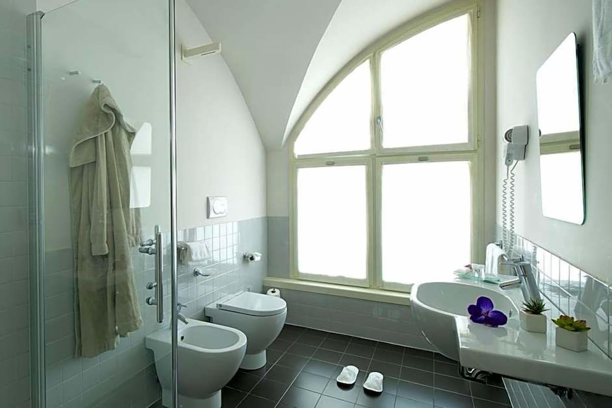Un baño moderno con cabina de ducha de vidrio, dos inodoros, un lavamanos y un amplio ventanal que proporciona luz natural.