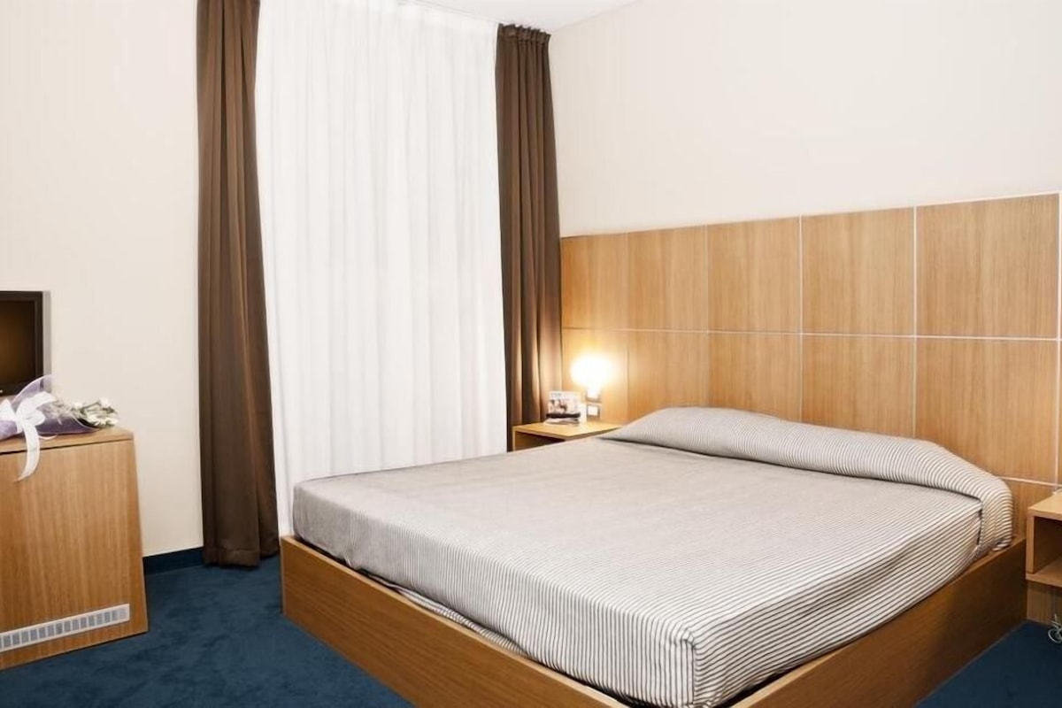 A hotel room with a wooden bed, a TV on a wooden dresser, and a nightstand with a lamp.