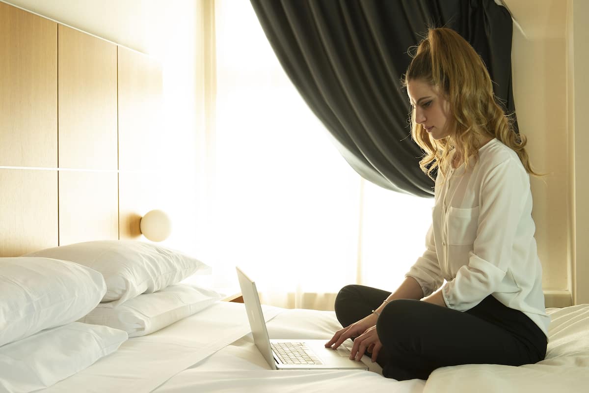 Eine Person sitzt auf einem Bett mit einem Laptop in einem Zimmer, das ein Holz-Kopfteil und ein großes Fenster mit Vorhängen hat.